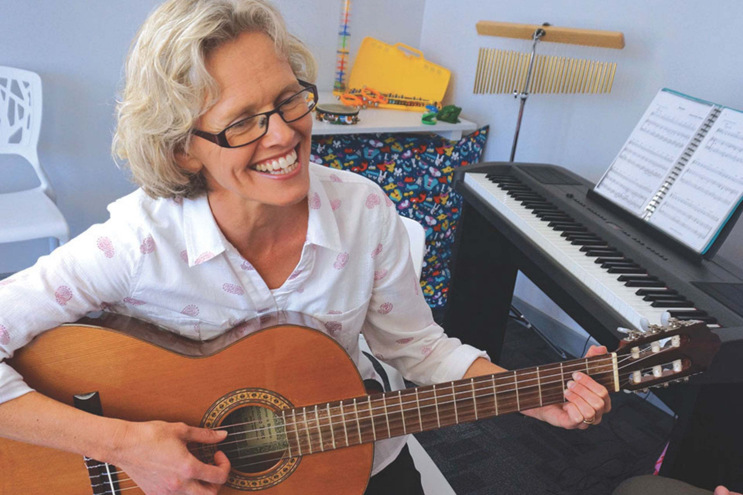 Susan Ashley-Brown - a woman with chin-length white hair wearing glasses and a white patterned shirt, playing a guitar and smiling